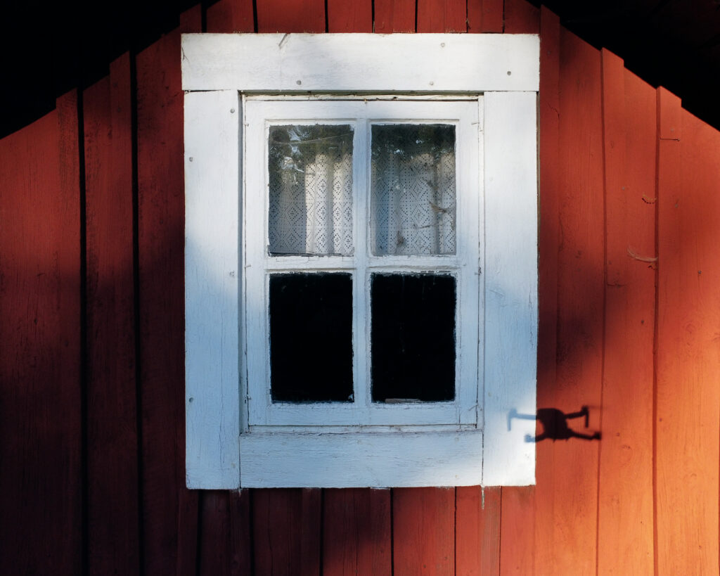 En rödmålad gavelhusvägg i trä. ett fönster mitt i bilden med vitt foder och skuggan av en drönare syns i det röda träet. En spetsgardin innanför det stängda fönstret och spindelväv syns både innanför och utanför fönstret.