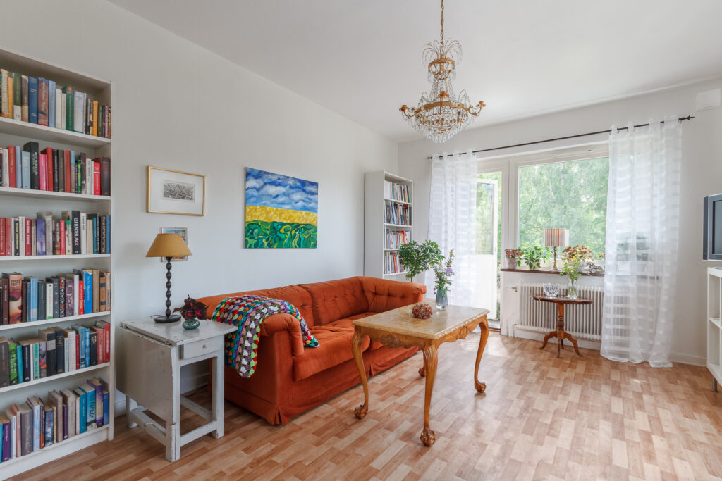 Interiörbild med orange soffa, soffbord med kurviga ben, vit bokhylla, en färgglad rutig, virkad filt och en öppen balkongdörr där man ser gröna sommarträd utanför. I taket en lampa med prismor. Väggarna är vitmålade och golvet träfärgat.