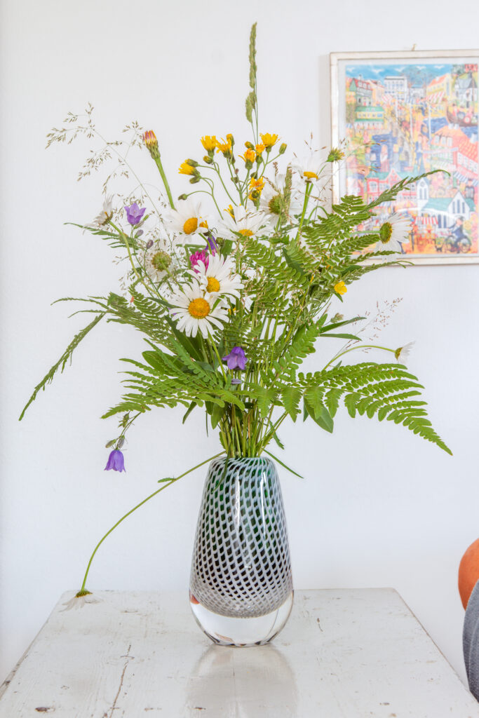 En grön glasvas med sommarblommor på ett repat ljust grått bord. Bakom en vitmålad vägg och en färgglad tavla av Gunilla Mann med Värnamomotiv.